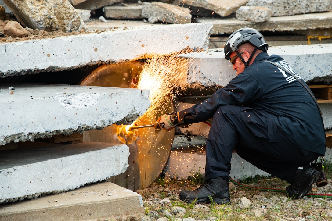 A firefighter wearing a blue uniform, helmet and protective glasses and gloves crouches on the ground and operates an acetylene torch on a metal drum located underneath stacked concrete. There are amber colored sparks flying off of where the torch and metal meet.