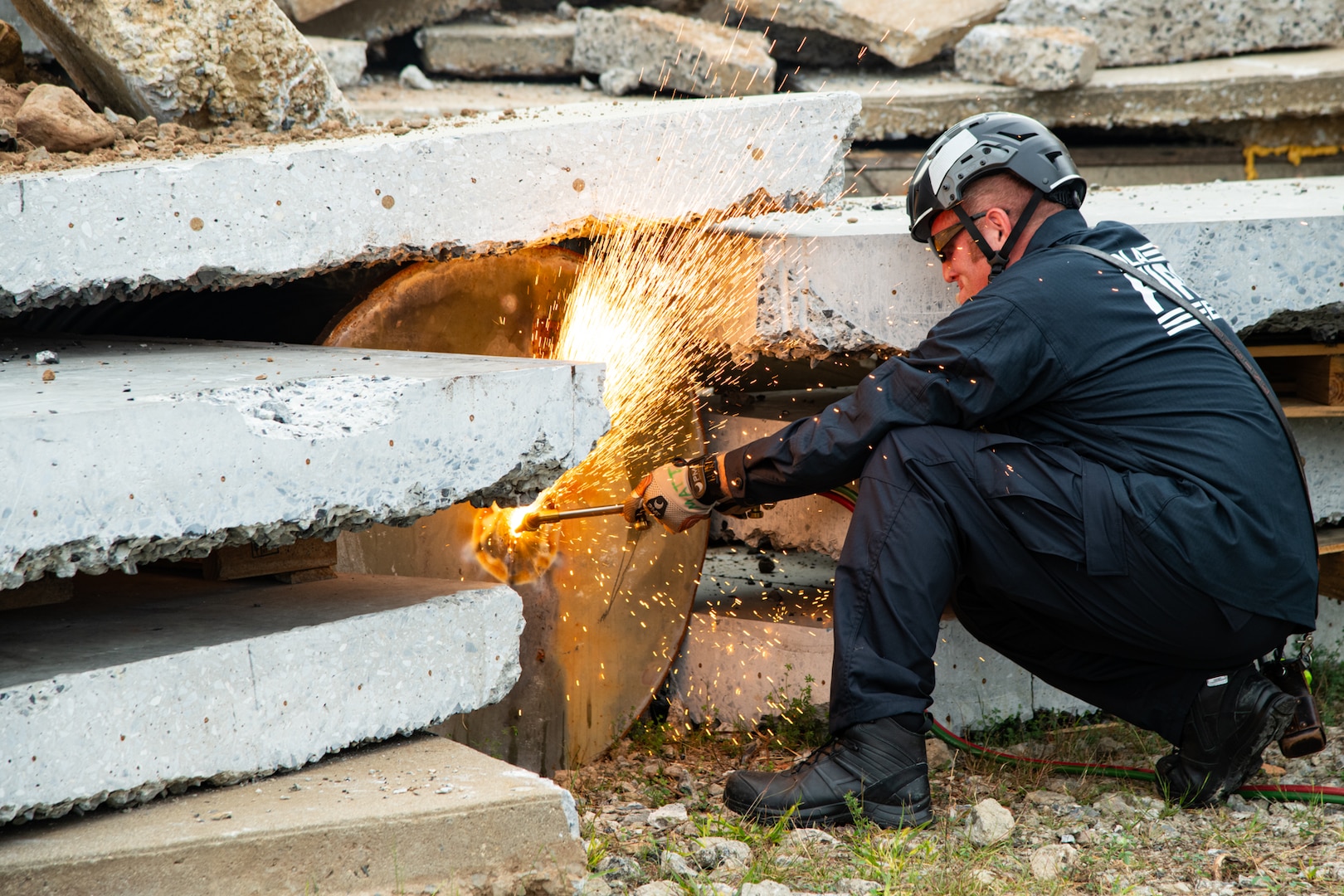 A firefighter wearing a blue uniform, helmet and protective glasses and gloves crouches on the ground and operates an acetylene torch on a metal drum located underneath stacked concrete. There are amber colored sparks flying off of where the torch and metal meet.
