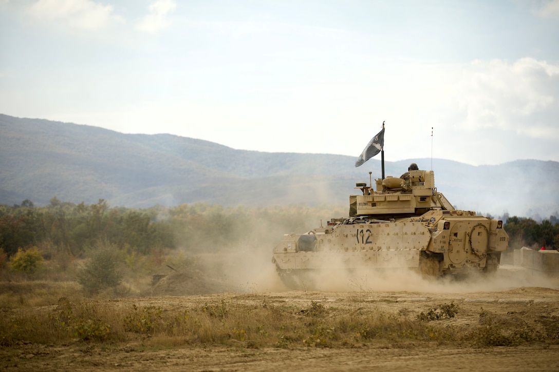 A soldier drives a military tank across a dusty field under a blue sky with mountains in the distance.