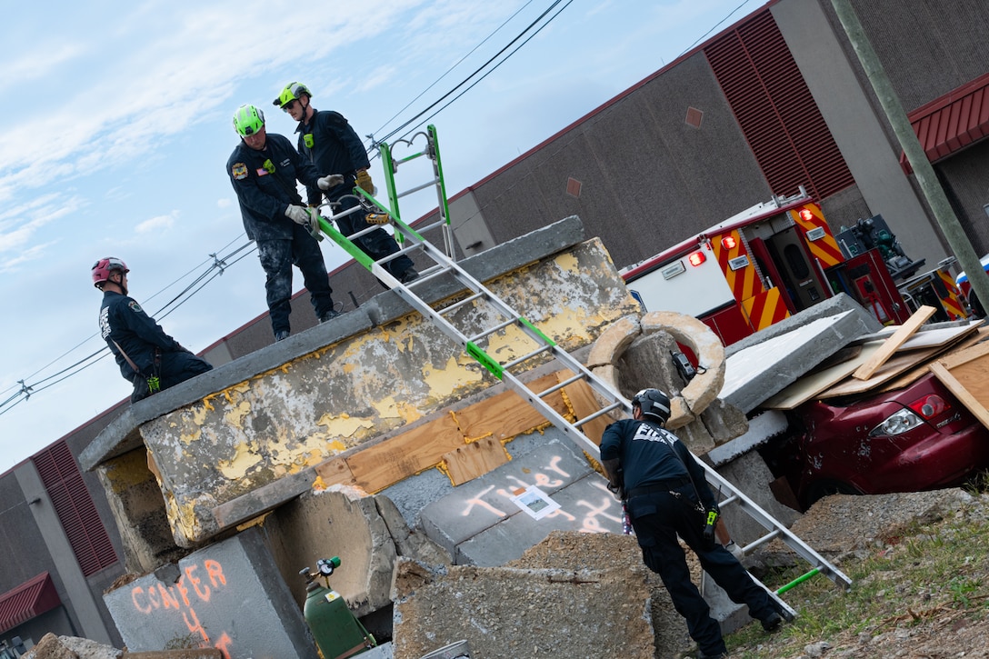 A group of firefighters wearing blue uniforms stand on top of a concrete structure outside. Another firefighter is standing on the ground propping a ladder up against the concrete structure.