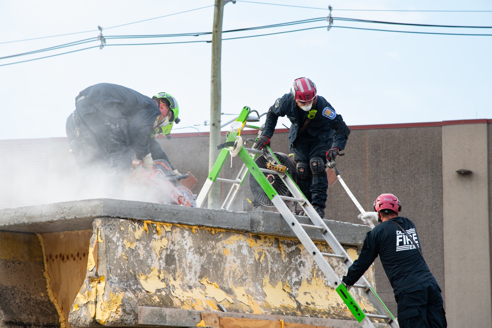 Three firefighters wearing blue uniforms, helmets and facemasks utilize circular saws on the top of a concrete structure. Another firefighter is climbing up a ladder toward them.