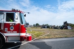 A red fire engine that says "DLA Fire & EMS" with the Defense Logistics Agency logo on it is in the foreground with firefighters working on concrete structures in the background.