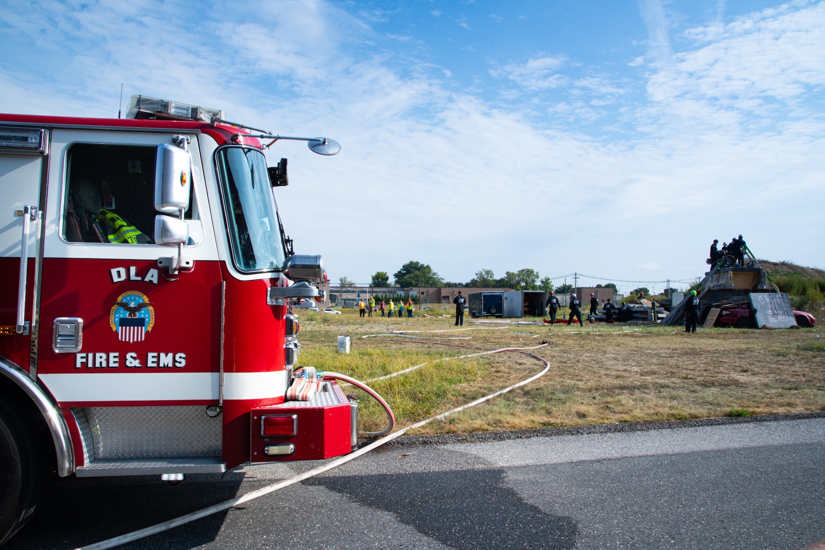 A red fire engine that says "DLA Fire & EMS" with the Defense Logistics Agency logo on it is in the foreground with firefighters working on concrete structures in the background.