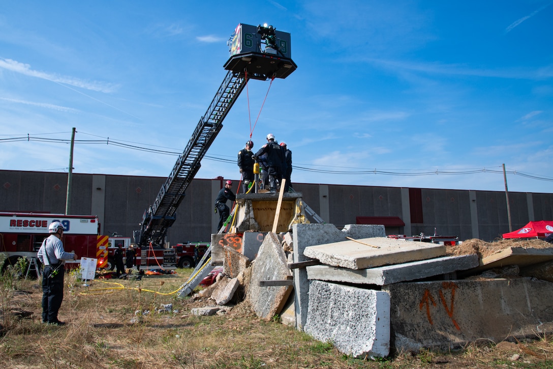 A group of firefighters wearing blue uniforms stand on top of a concrete structure outside. There is a fire engine parked behind the structure with an aerial platform extending over top of the concrete structure.
