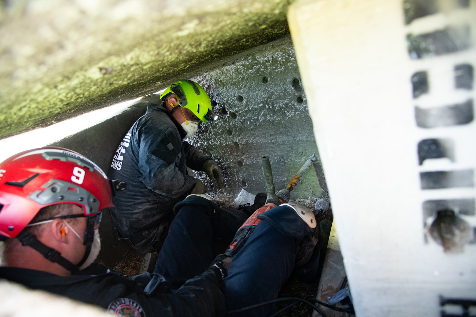 Two firefighters wearing blue uniforms, helmets and facemasks, drill into a concrete structure within a confined space.