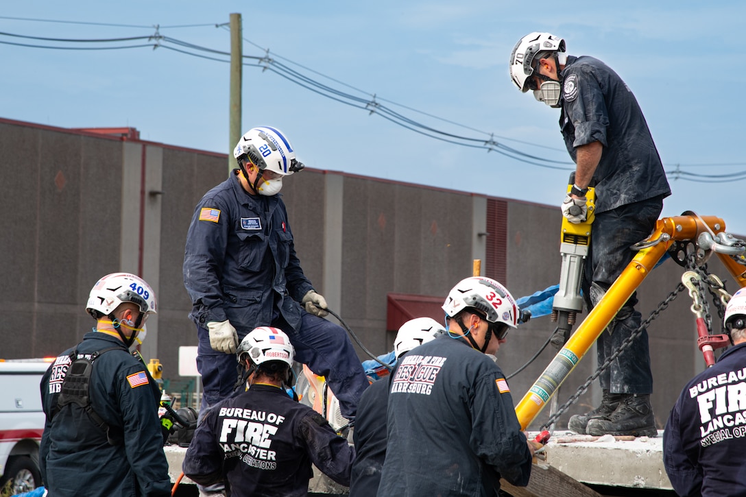 A group of seven firefighters all wearing blue uniforms stand around a concrete structure. One firefighter operating a jackhammer on the top of the structure.