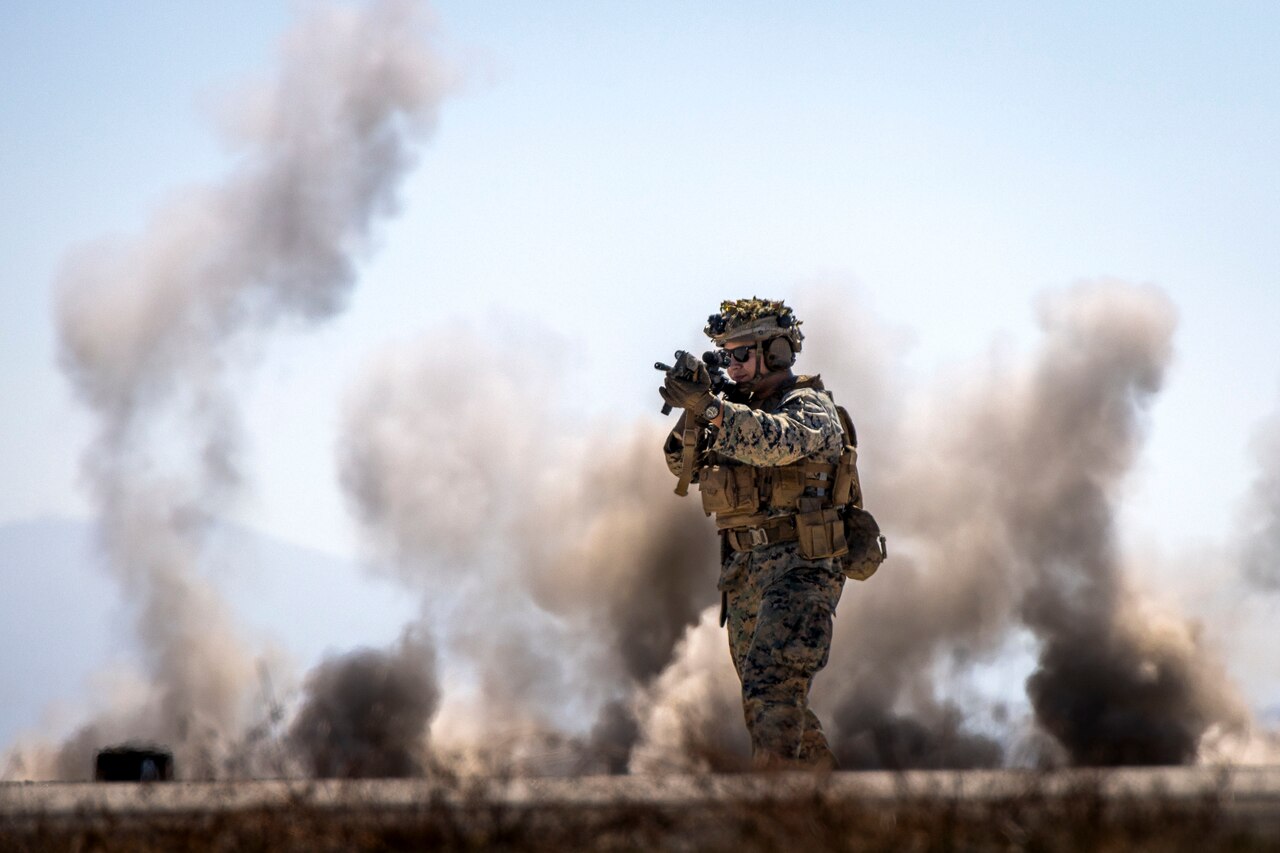 A Marine in tactical gear stands in front of columns of smoke during the day and aims a service weapon.