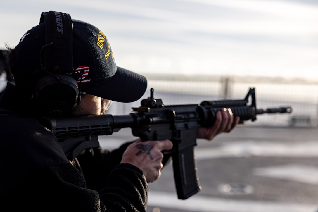 A sailor in a baseball cap aims a service weapon to the right while standing on a ship deck, as seen from behind.