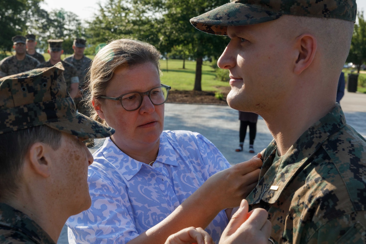 A person in a camouflage military uniform and a person in casual attire pin a military rank on the collar of another person wearing a military uniform outside.