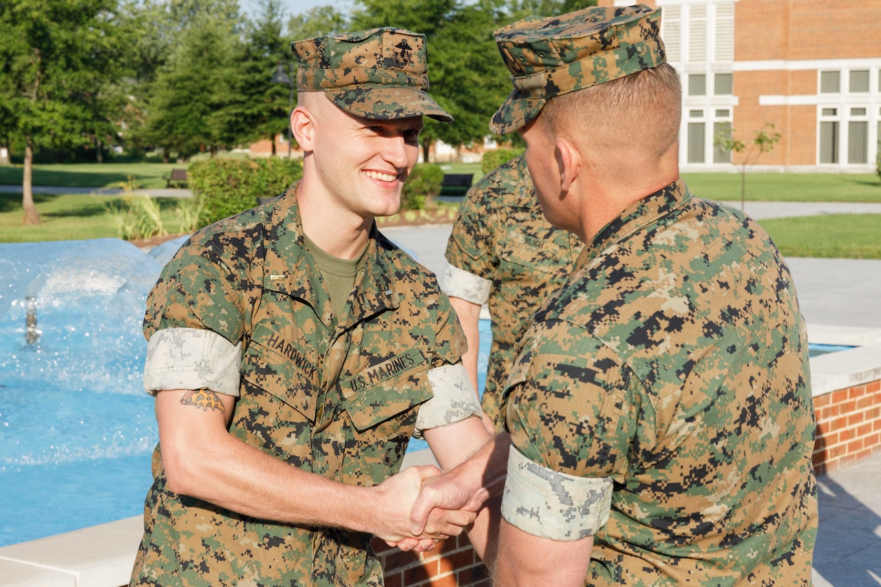 Two people wearing camouflage military uniforms shake hands outside.
