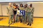 Members of Walter Reed/Navy Medicine Readiness and Training Command (NMRTC) Health Promotion and Wellness Committee show off the U.S. Navy Surgeon General's Blue H flag and ribbon Gold Star awarded by the U.S. Navy SG for health promotion and wellness programs and activities held at Walter Reed during 2024 promoting responsible drinking, healthy eating, active living, psychological wellness, sexual heath, tobacco-free living and weight management.
