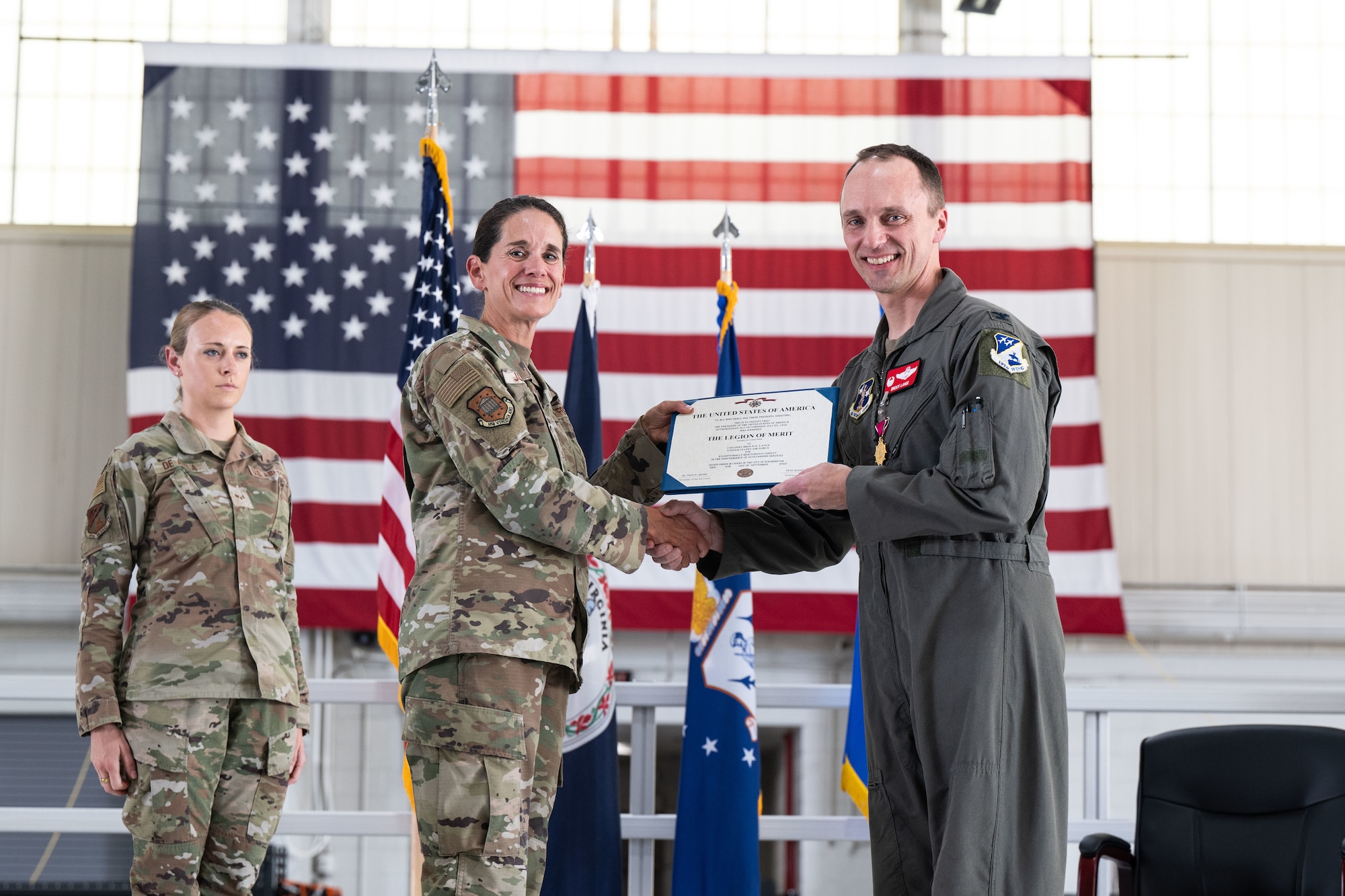 A U.S. military officer in a flight suit receives the Legion of Merit award certificate from a female officer in a camouflage uniform during a formal ceremony. Both are smiling and shaking hands, with a large American flag and several military flags in the background. Another service member in camouflage stands at attention to the side.