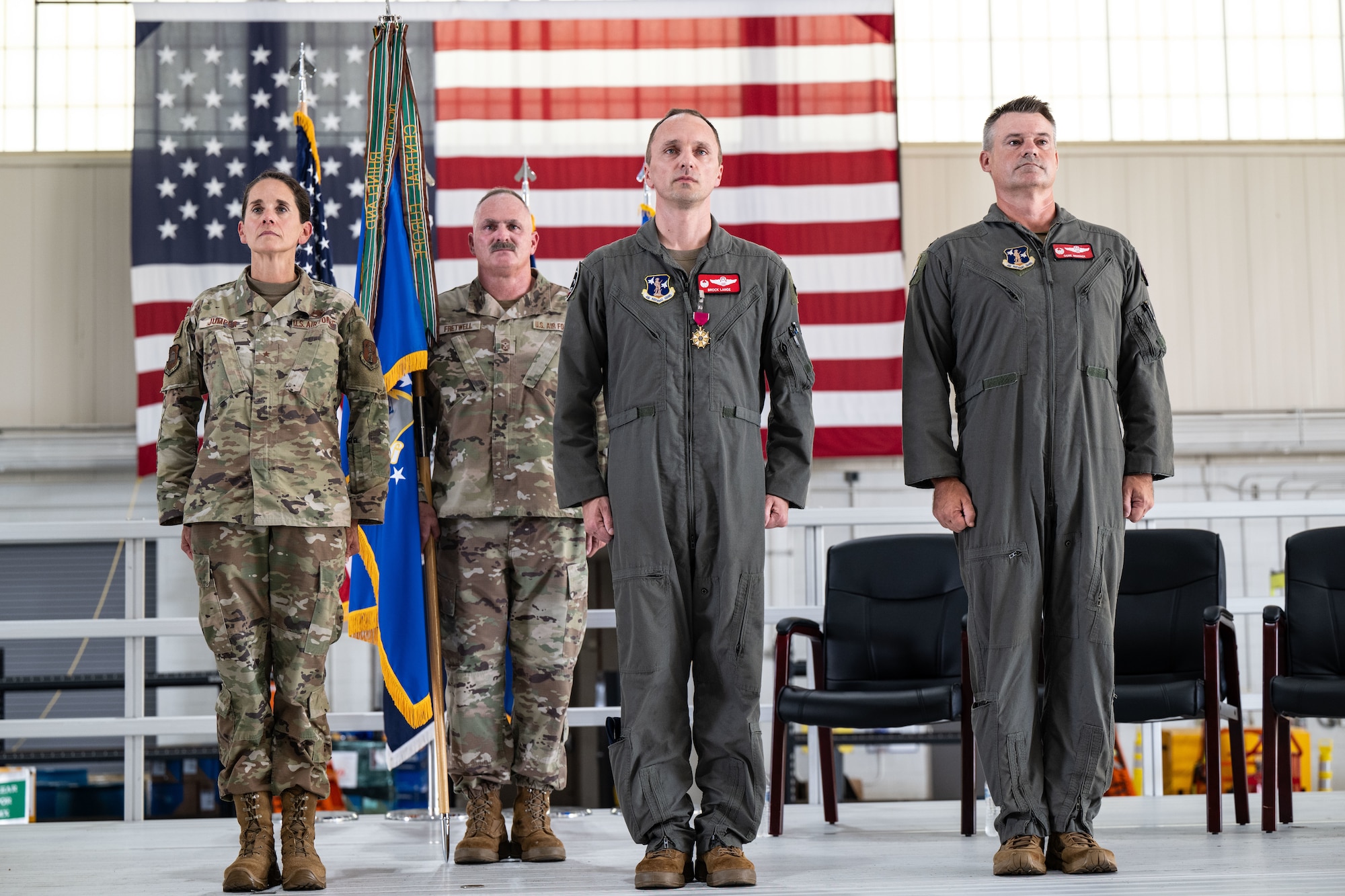 Four U.S. service members stand at attention during a formal military ceremony in front of a large American flag. Two are in camouflage uniforms and two are in green flight suits. One officer in a flight suit wears a medal on his chest. A ceremonial flag is held between the two individuals in camouflage.
