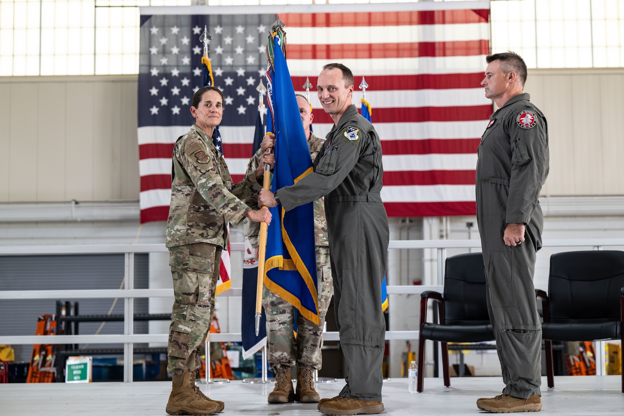 A U.S. military change of command ceremony is held indoors, in front of a large American flag. A female officer in a camouflage uniform hands a unit guidon (flag) to a male officer in a green flight suit, symbolizing the transfer of command. Another service member stands behind them holding the flagpole, and a third officer in a flight suit stands to the side, observing the ceremony.