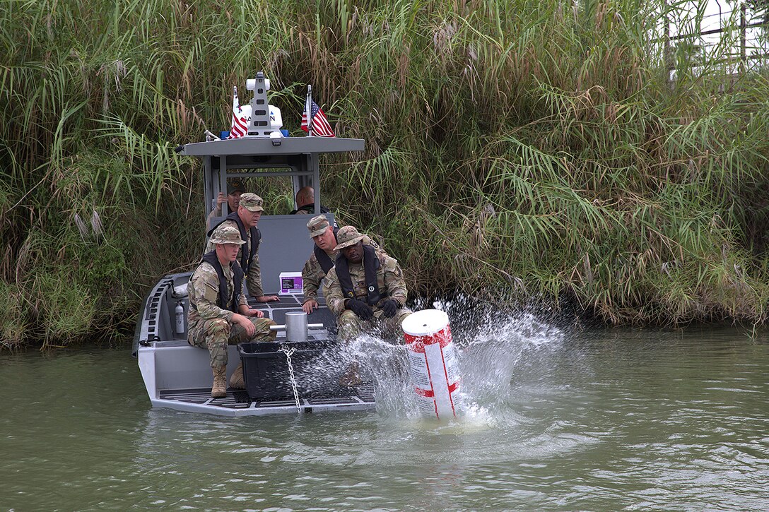 Soldiers in camo aboard a small motor boat in a river near shoreline watch as a buoy splashes into the water.