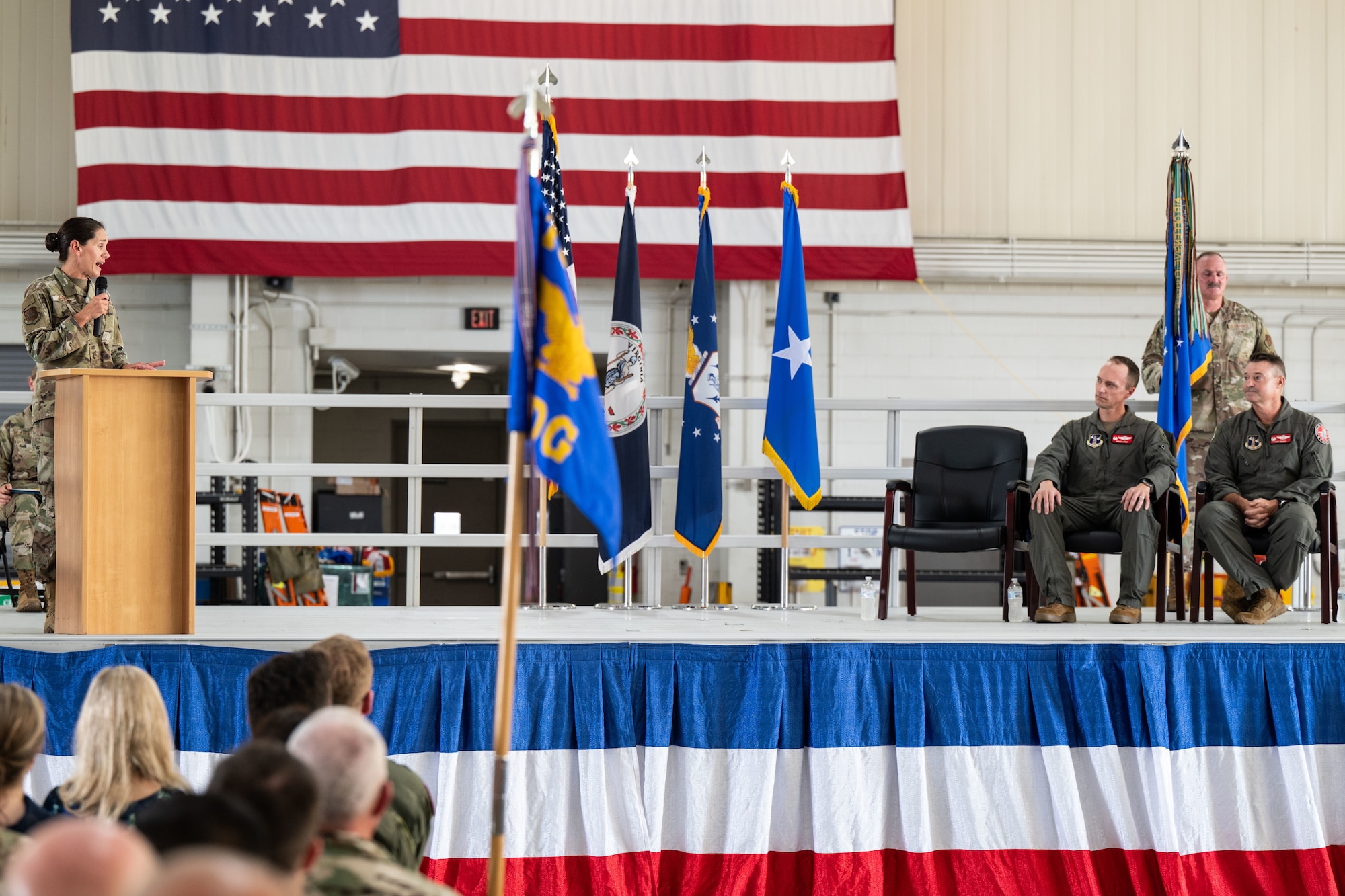 A military ceremony inside a hangar, with a large American flag hanging on the wall. On stage, two men in flight suits sit in chairs while a man in camouflage uniform stands behind them holding a guidon. To the left, a woman in camouflage uniform speaks at a wooden podium. Several flags are displayed across the stage, and an audience watches from the foreground.