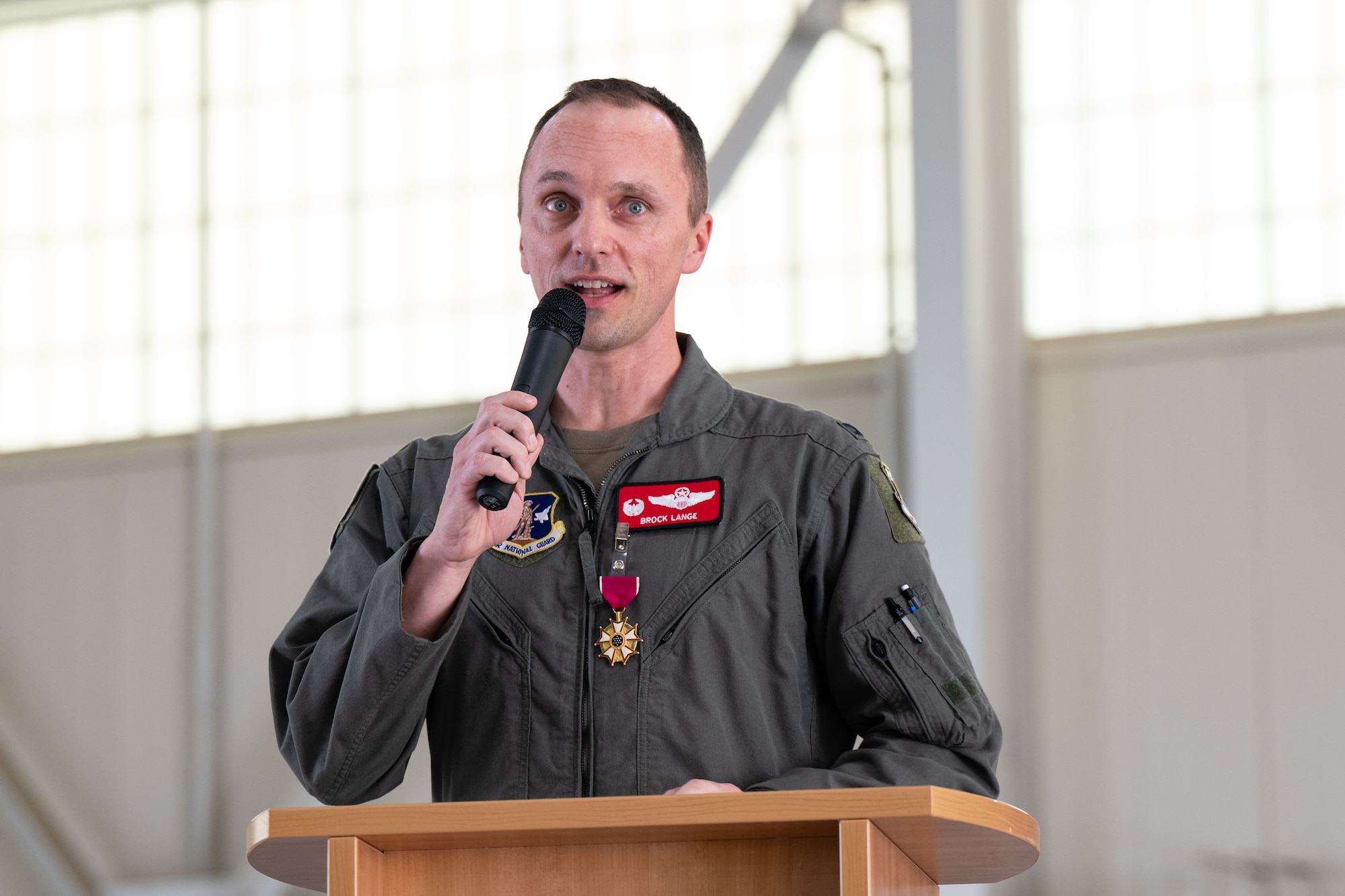 A U.S. Air National Guard member in uniform, Col. Brock Lange, speaks into a microphone while standing at a wooden podium. He is wearing a flight suit with patches and a Legion of Merit medal pinned to his chest, inside a large, well-lit hangar.