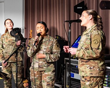 Three Army soldiers in combat uniforms are performing music into microphones, and one is holding a black violin.