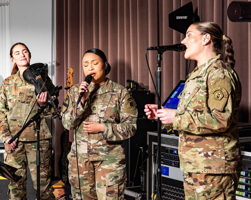 Three Army soldiers in combat uniforms are performing music into microphones, and one is holding a black violin.