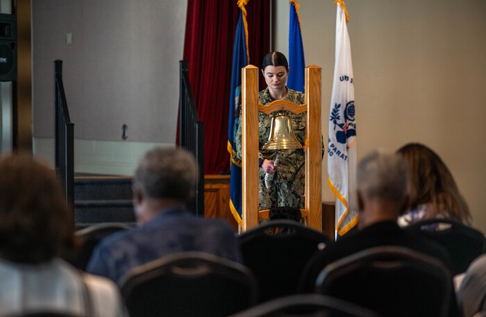 A female Navy Officer rings a large bell in front of a crowd
