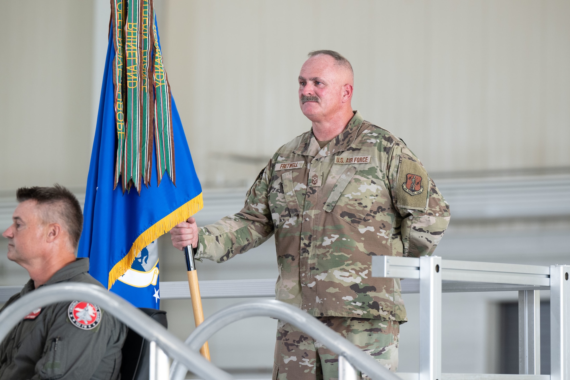 Chief Master Sgt. Sean Fretwell, Virginia Air National Guard, stands in camouflage uniform holding a blue guidon decorated with unit streamers during a ceremony inside a hangar. In the foreground, Col. Andrew Weidner is seated in a flight suit with a Virginia Air National Guard patch.