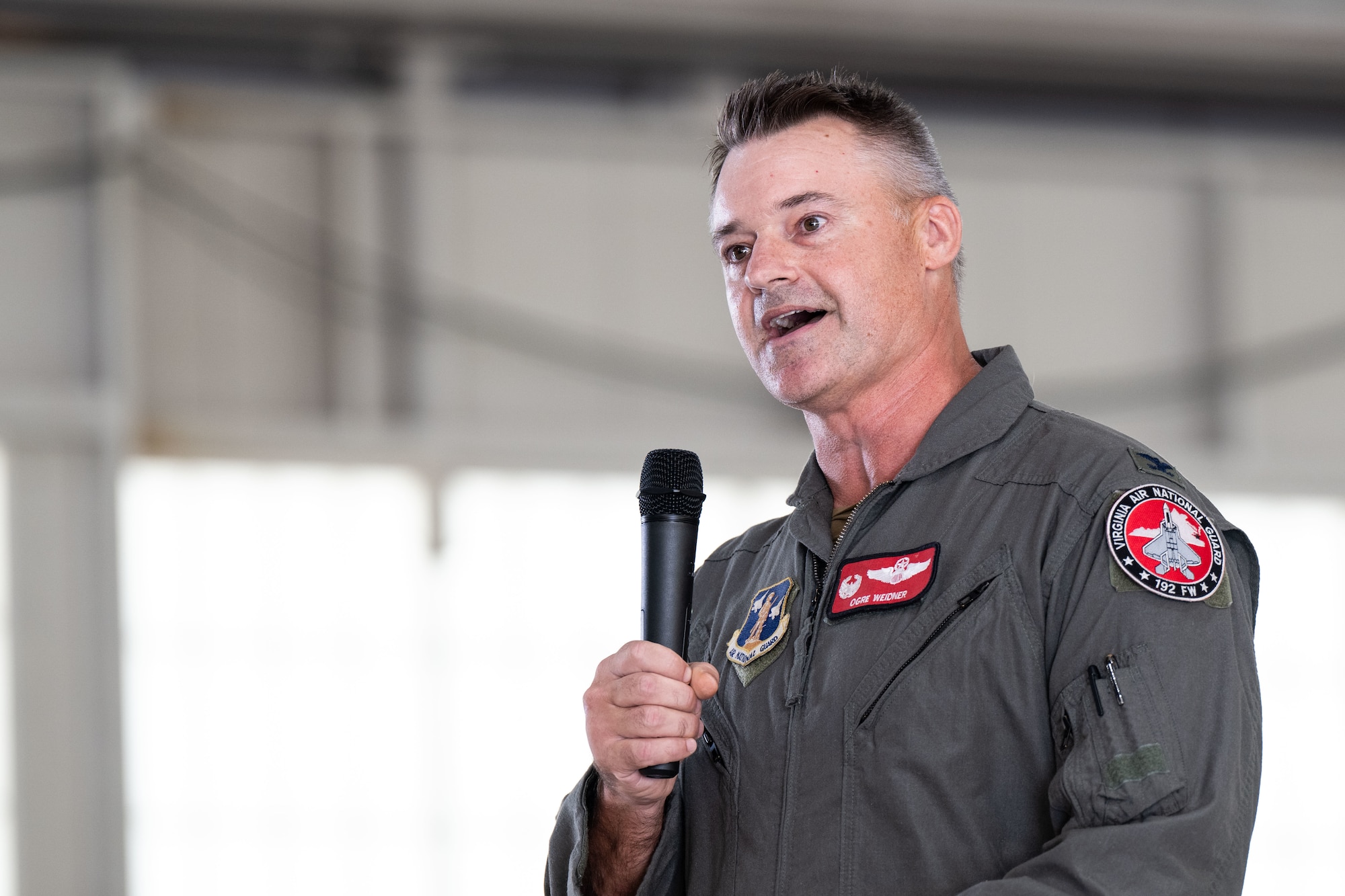 Col. Andrew Weidner, a U.S. Air National Guard member, speaks into a microphone during a ceremony inside a hangar. He is wearing a flight suit with patches, including one that reads "Ogre Weidner."