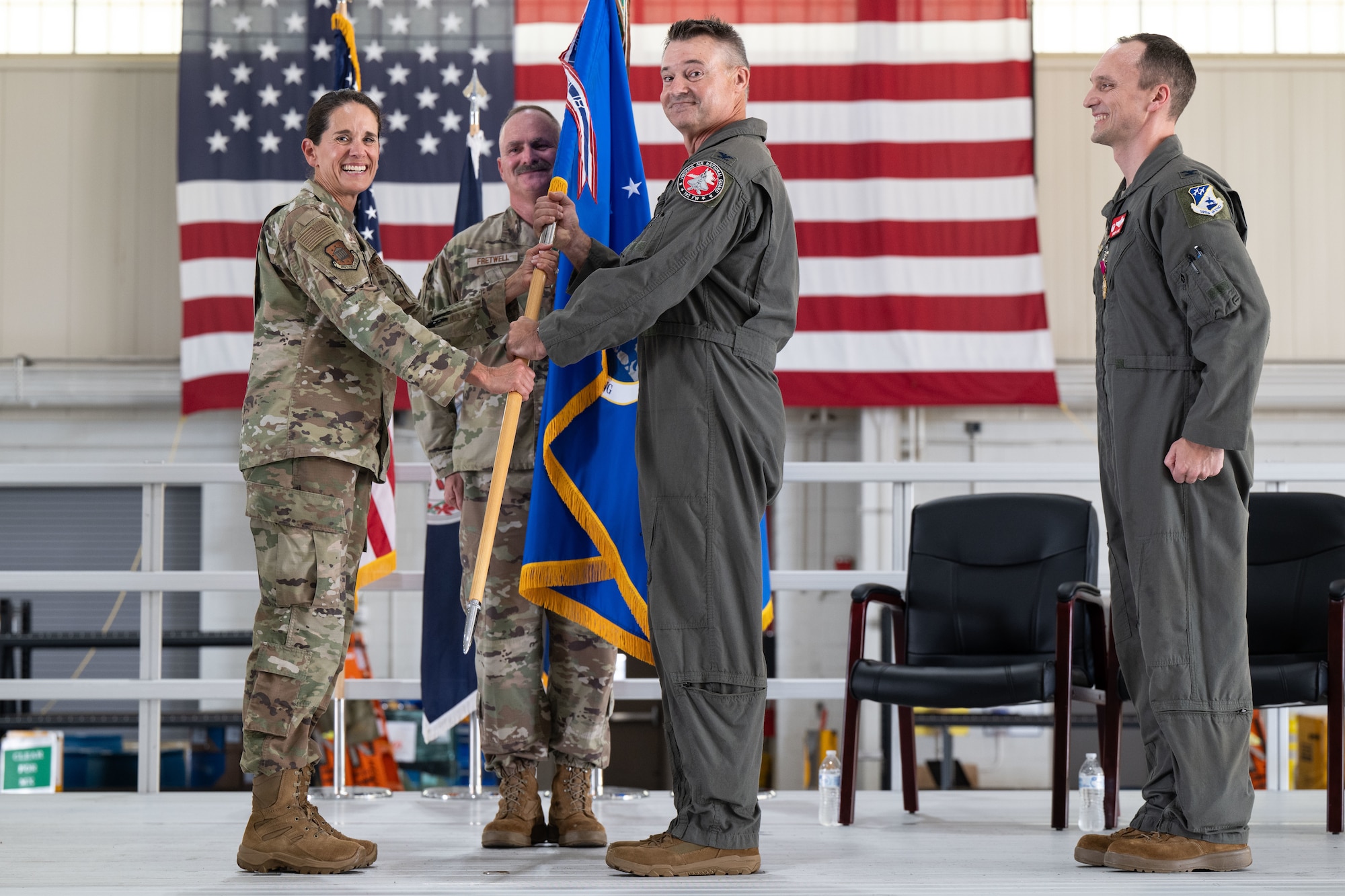A U.S. military change of command ceremony takes place indoors with an American flag in the background. A woman in Air Force camouflage uniform hands a unit guidon (flag) to a man in a green flight suit, symbolizing the transfer of command. Two other men, also in military uniforms, observe the ceremony and smile.
