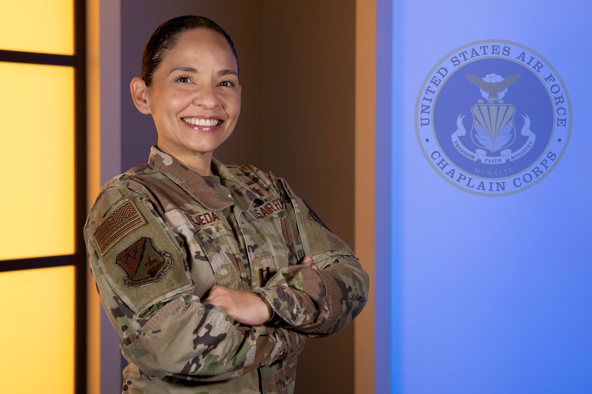 A smiling U.S. Air National Guard member, Chaplain KMarie Tejeda, in camouflage uniform stands with arms crossed in front of a blue background displaying the seal of the United States Air Force Chaplain Corps. She wears the rank of captain on her chest. A warm yellow light panel is visible on the left side of the image.