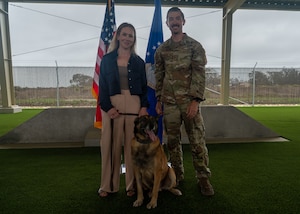 Two people stand behind a dog for a portrait photo outside. They smile for the camera.