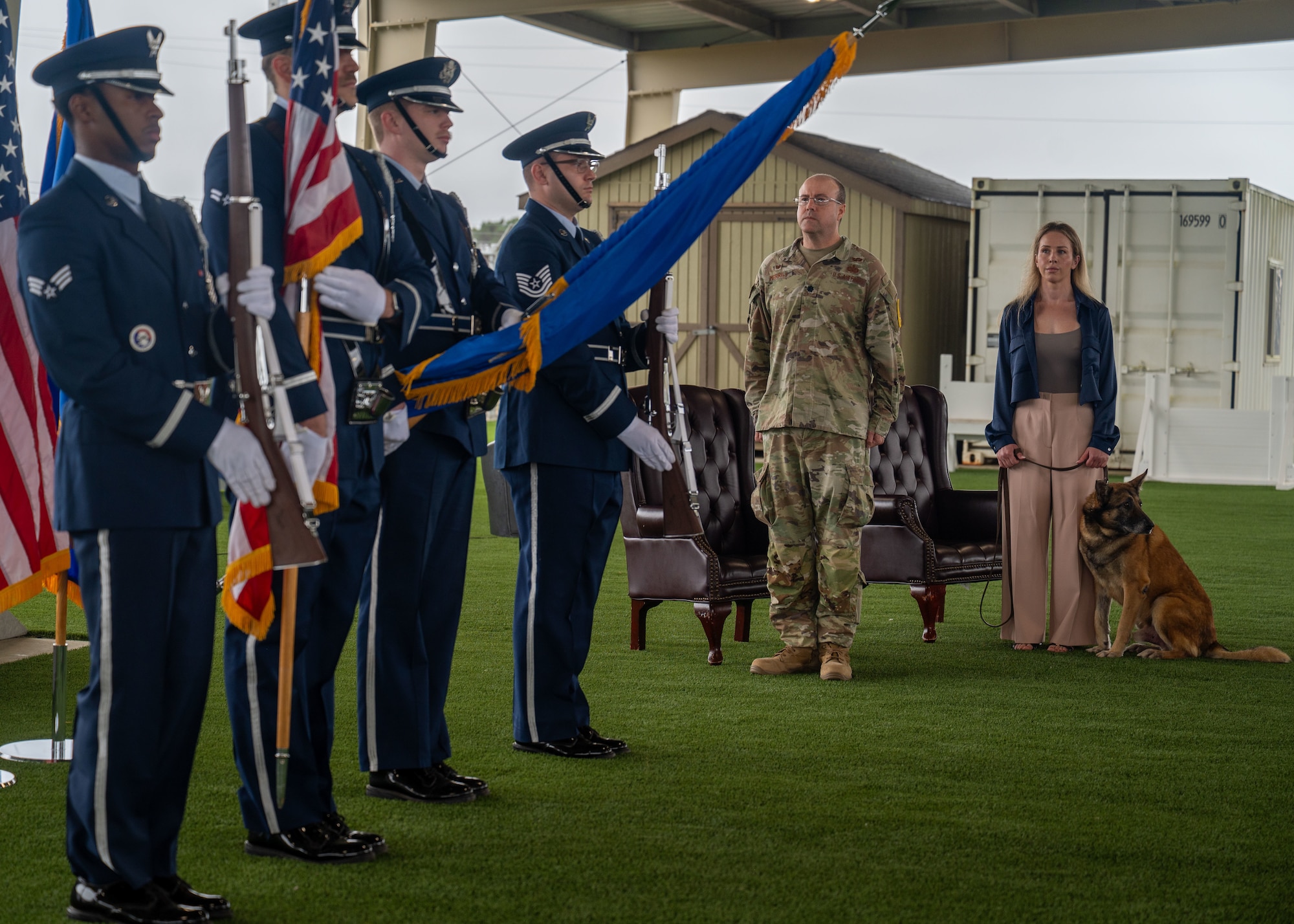 Four honor guard members stand for the presentation of the colors. A colonel and civilian stands aside, holding a dog on a leash.