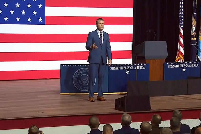 Secretary of War Pete Hegseth speaks on a stage with a U.S. flag in the background.