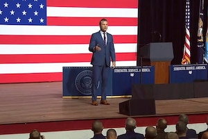 Secretary of War Pete Hegseth speaks on a stage with a U.S. flag in the background.