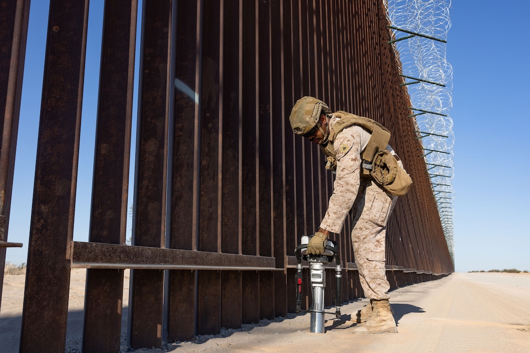 A Marine handles a metal device lodged in flat, arid ground next to a tall brown wall.