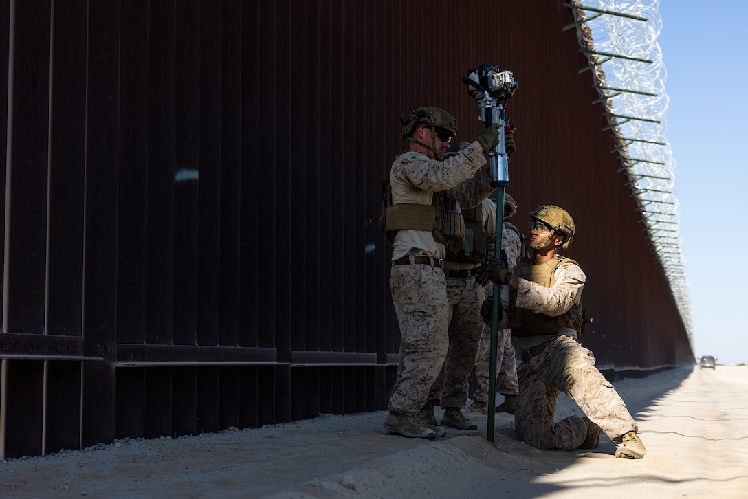 Several Marines handle a metal signpost by a tall brown wall along desert terrain.