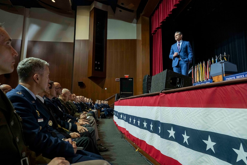 A person in a blue suit stands on stage and speaks to seated military officials, as seen from their seats.