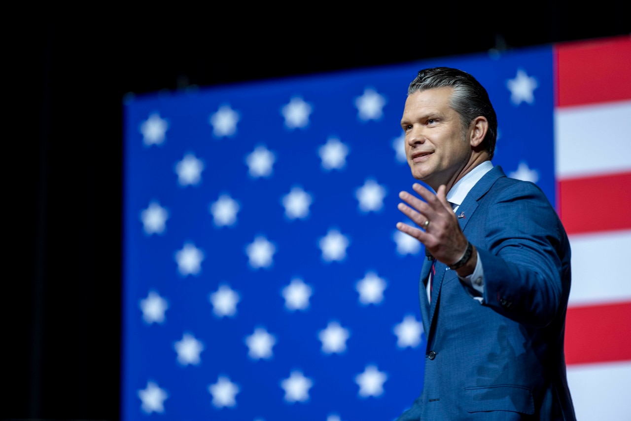 A person in a blue suit gestures while standing in front of a large American flag on a stage.