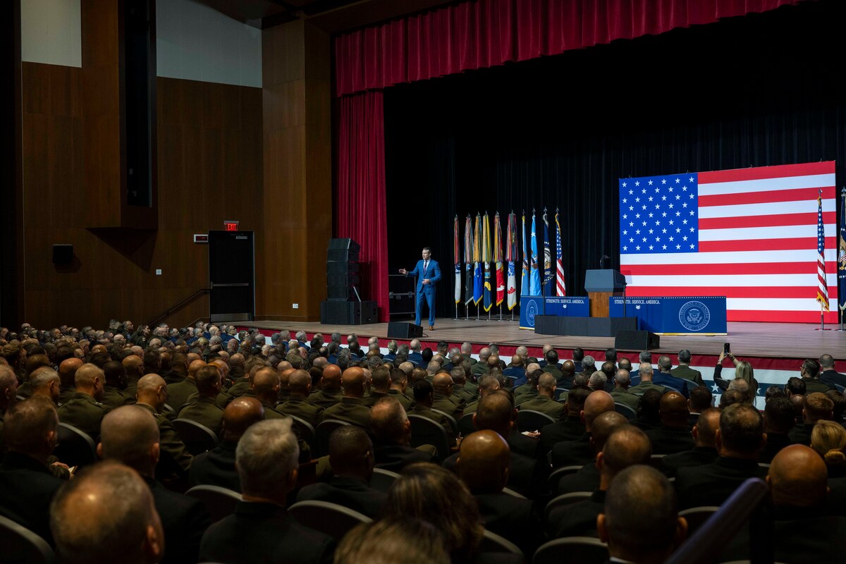 A person in a suit talks on stage to people seated in an auditorium with a large American flag behind.