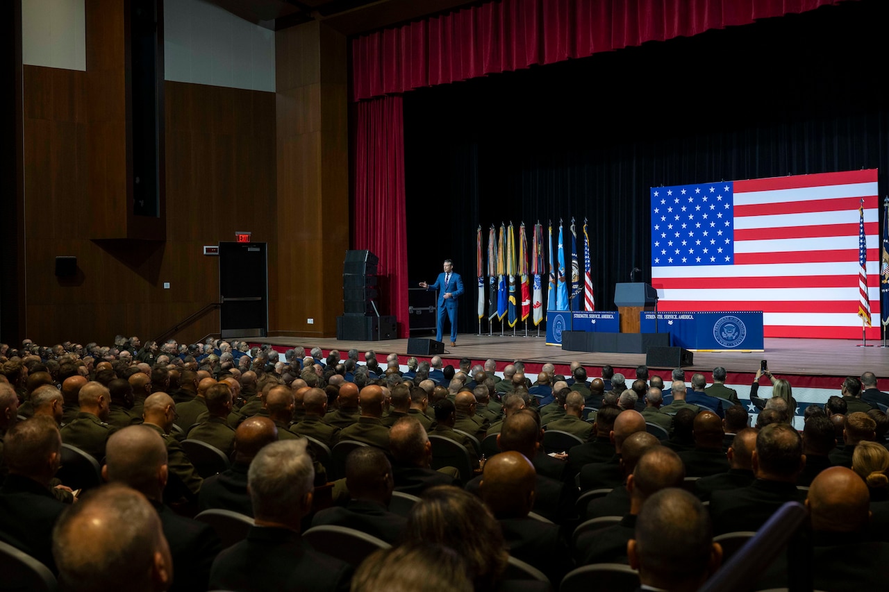 A person in a suit talks on stage to people seated in an auditorium with a large American flag behind.