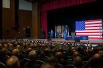 A person in a suit talks on stage to people seated in an auditorium with a large American flag behind.