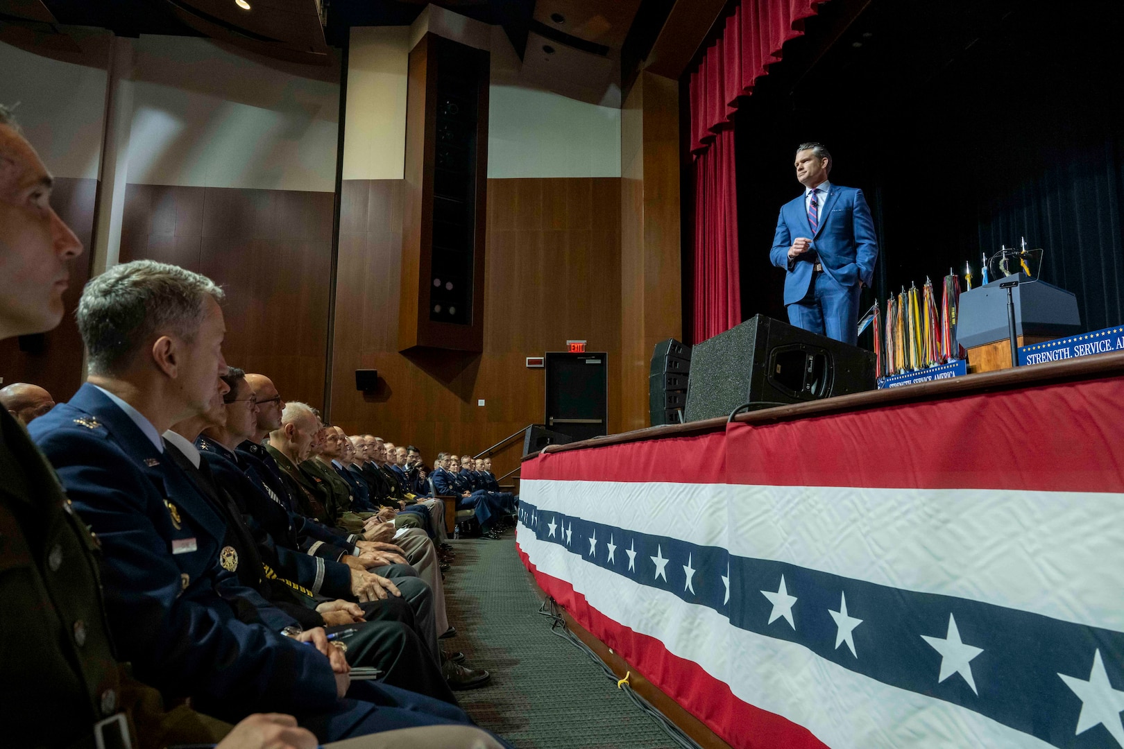 A person in a blue suit stands on stage and speaks to seated military officials, as seen from their seats.