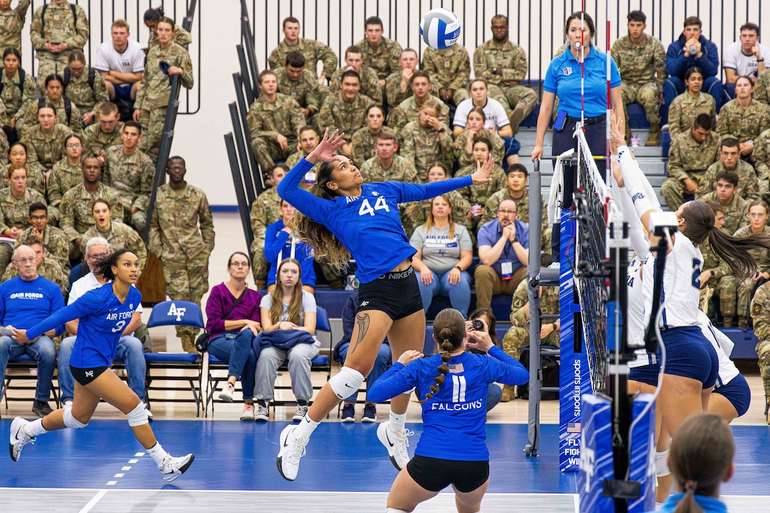A person jumps and prepares to hit a volleyball over a net as other players race around them, and people sitting in bleachers watch from behind.