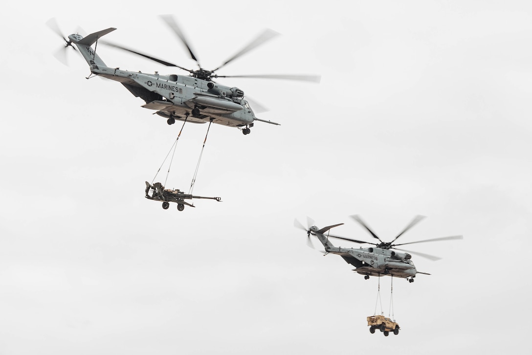 A vehicle hangs from a helicopter in flight next to another helicopter towing large artillery against a gray sky.