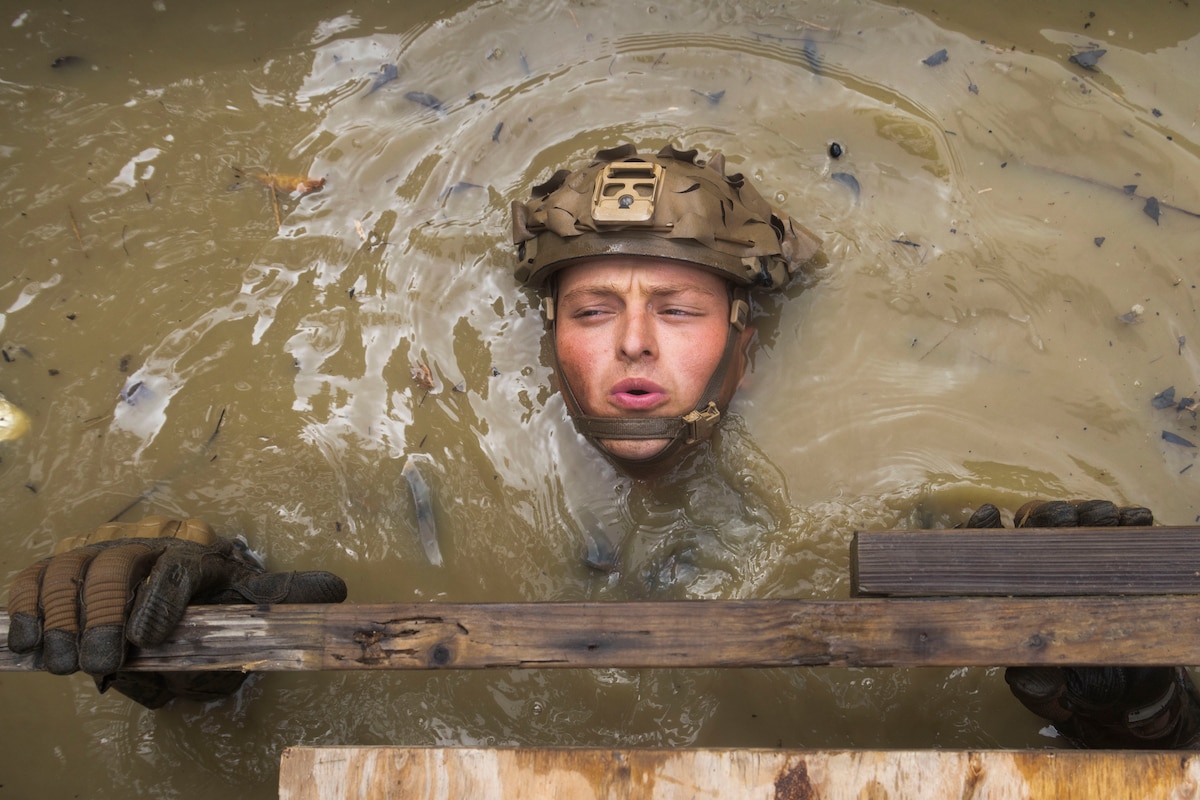 A submerged Marine holds their head above brown water while their gloved hands hold a piece of wood.