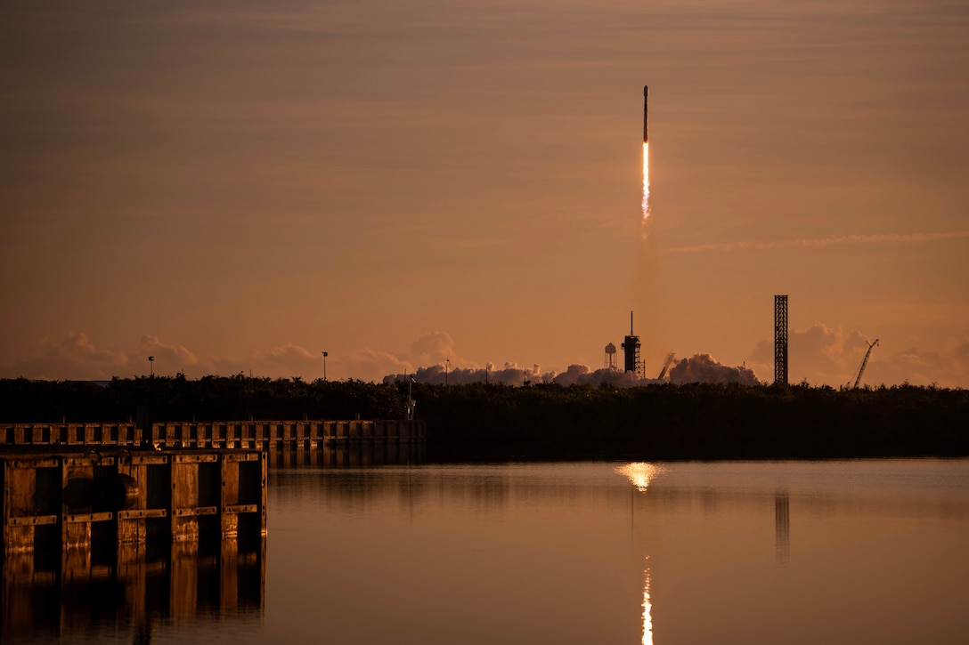 A rocket launches into a deep orange sky over a body of water with wooden docks, leaving smoke and a fiery trail behind.