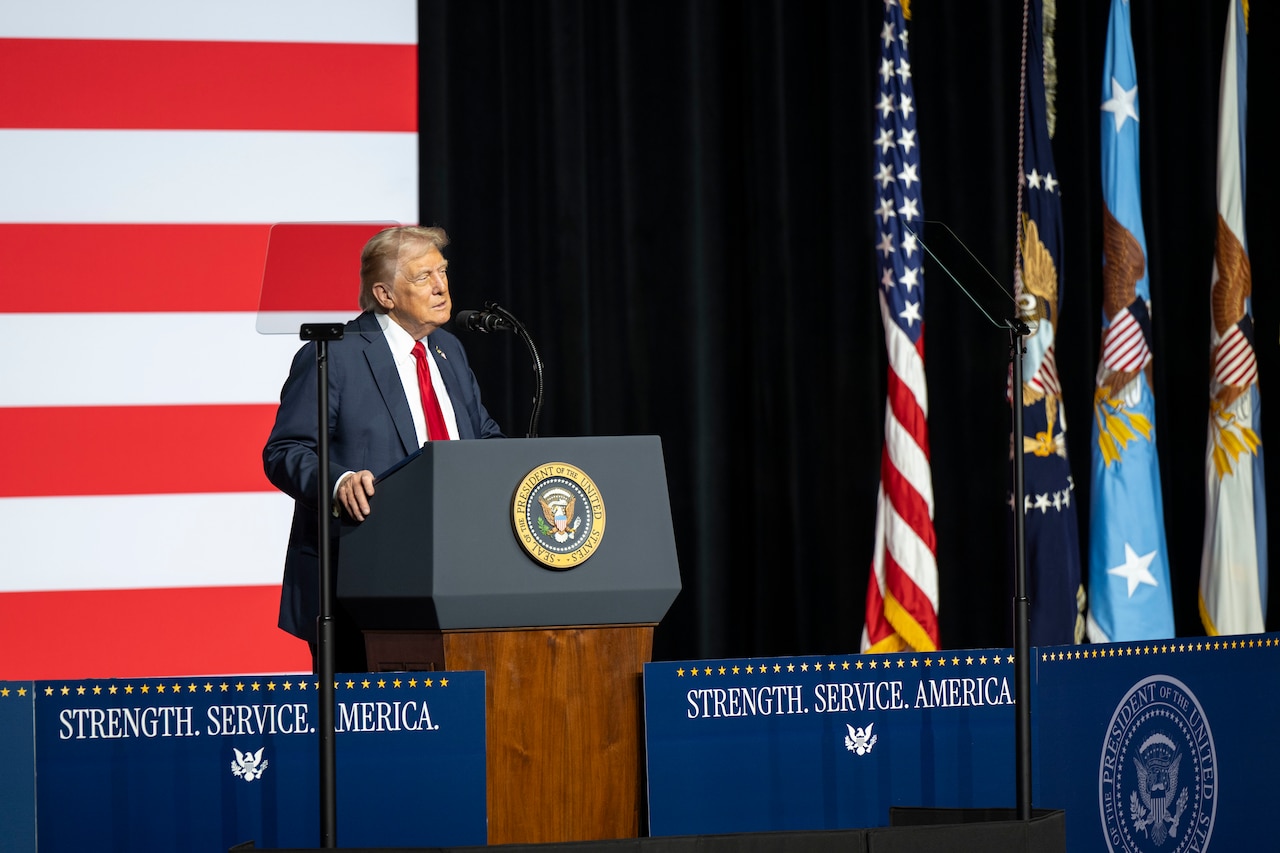 A person in a business suit stands behind a lectern while giving a speech. Behind him is a large American flag.