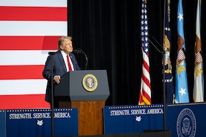 A person in a business suit stands behind a lectern while giving a speech. Behind him is a large American flag.