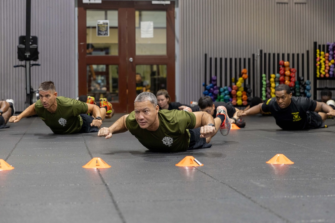 Soldiers laying on the floor of a gym hold their arms out, with their hands in fists, behind small orange cones and in front of colorful weights and a door.
