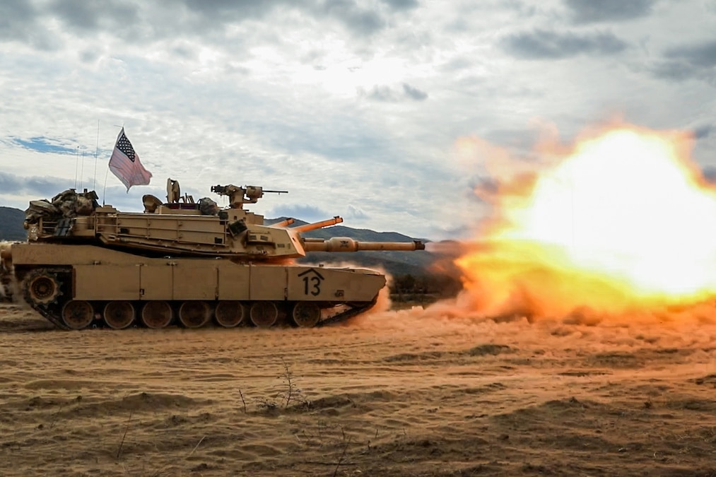 A military tank drives across a sandy field under a cloudy sky and fires, leaving a burst of orange flames in front.