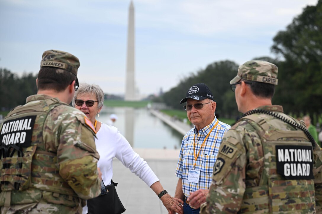 Two guardsmen stand facing two people in sunglasses during the day with the Washington Monument and the Reflecting Pool in the distance.