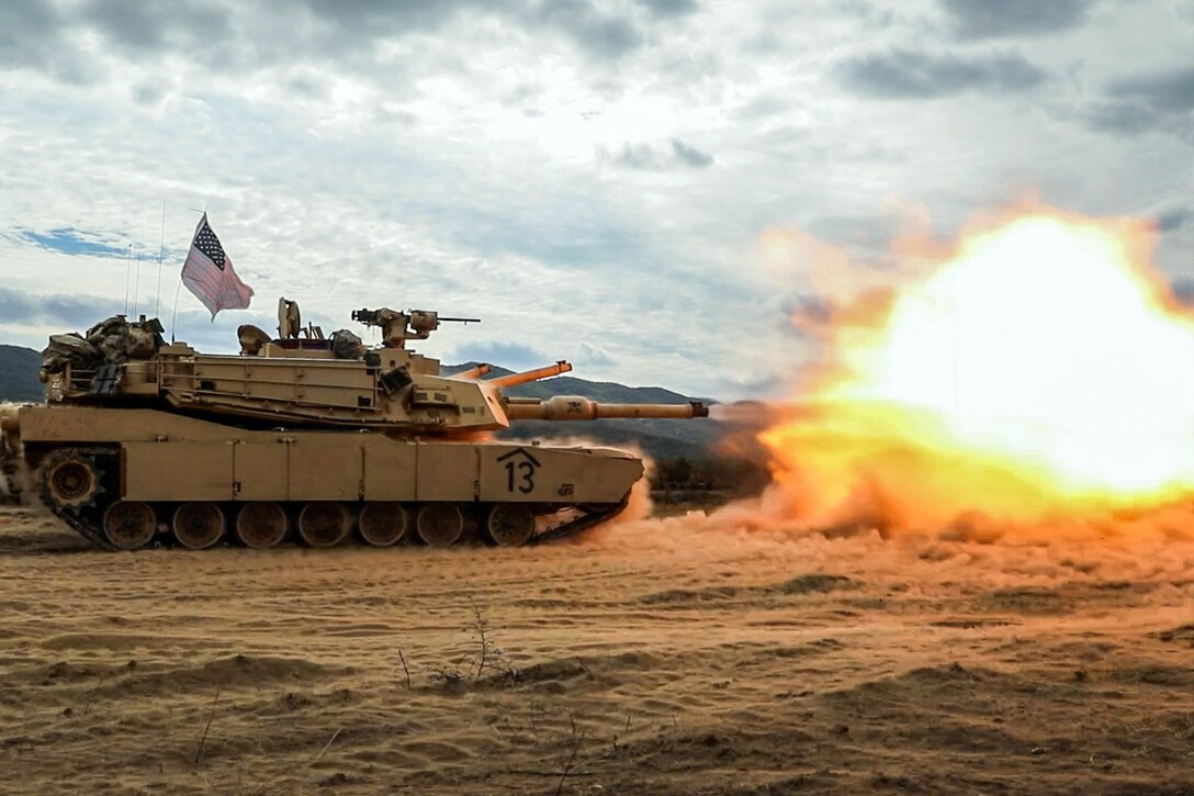 A military tank drives across a sandy field under a cloudy sky and fires, leaving a burst of orange flames in front.
